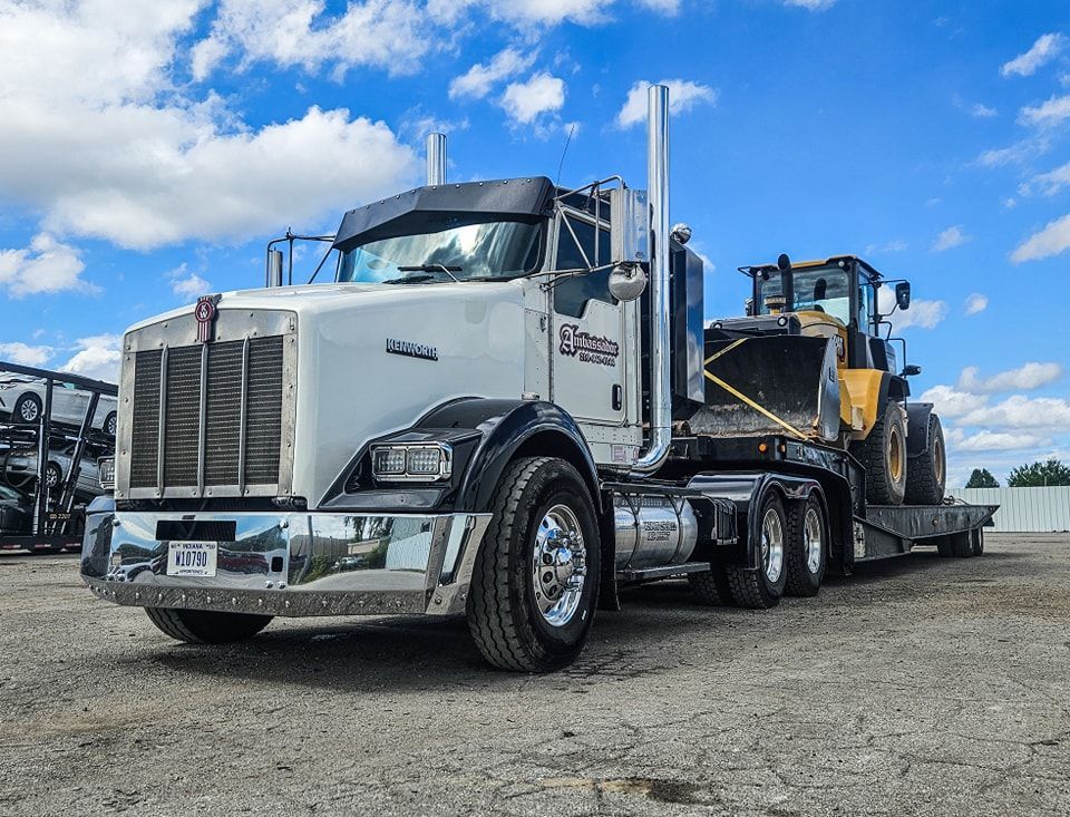 White Kenworth semi-truck with a flatbed trailer transporting a yellow front-end loader against a blue sky.