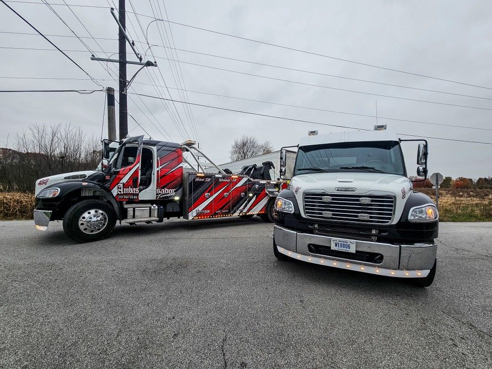 Tow truck towing a white semi-truck on a gray, overcast day; asphalt road, utility poles, and field in the background.