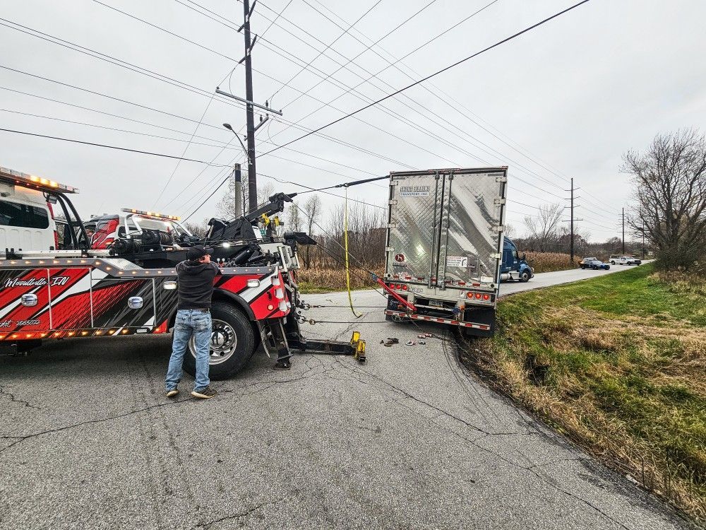 A tow truck operator pulls a semi-trailer from a ditch next to a road, under power lines.