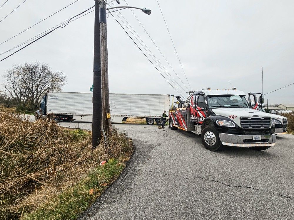 A tow truck attempts to free a semi-truck from a narrow road; gray sky and dry roadside vegetation.