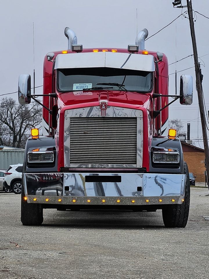 Red Kenworth semi-truck, front view, chrome grill and bumper, headlights on, parked outside.