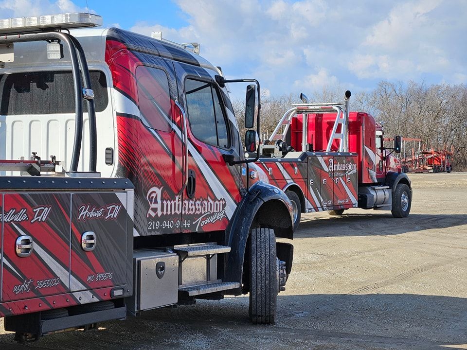 Red and black Ambassador tow truck parked in a gravel lot under a partly cloudy sky.