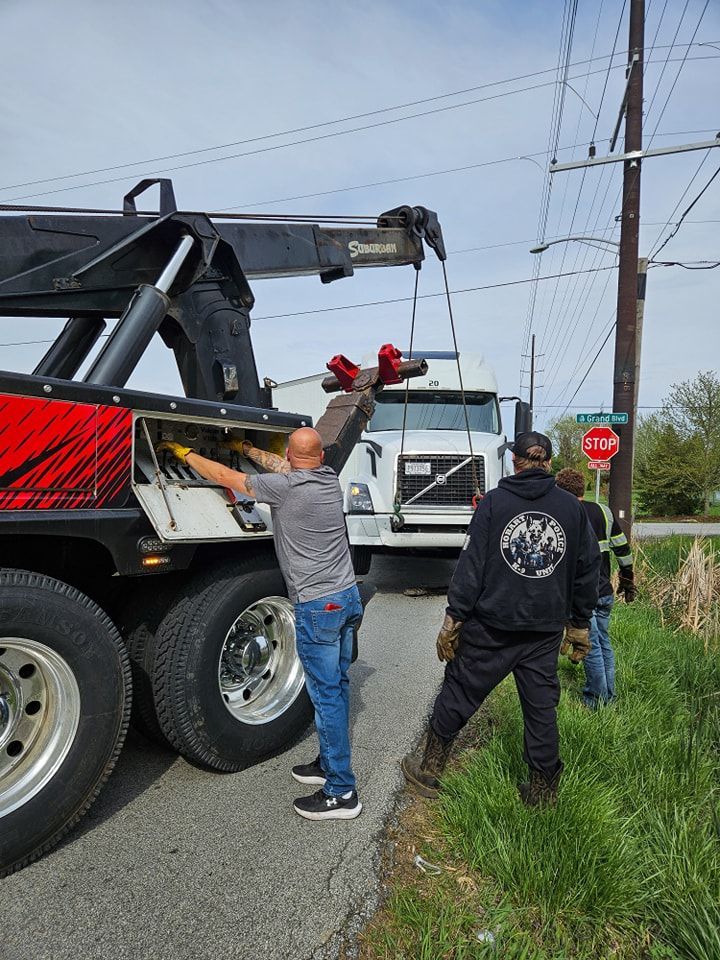 Tow truck operator prepares to tow a white semi-truck on a road. Two other people stand nearby.