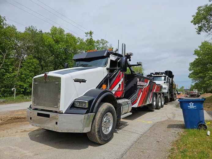 White and red tow truck on a paved road, parked next to a blue trash can and a second tow truck.