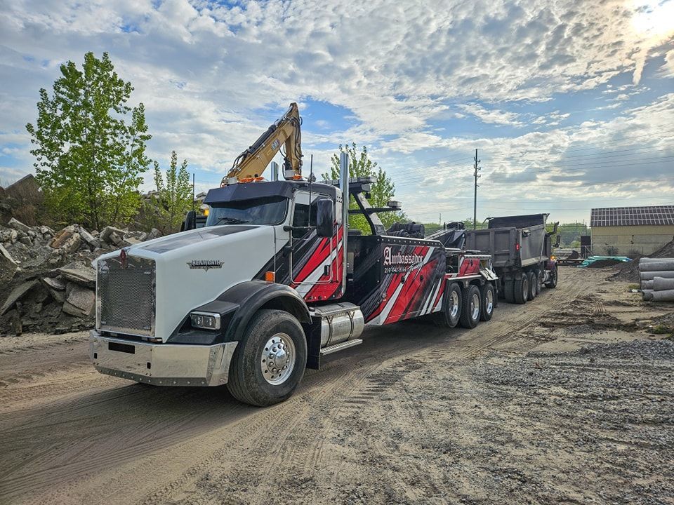 Tow truck with excavator on its bed, hauling a dump truck trailer. Bright sky, outdoor construction site.