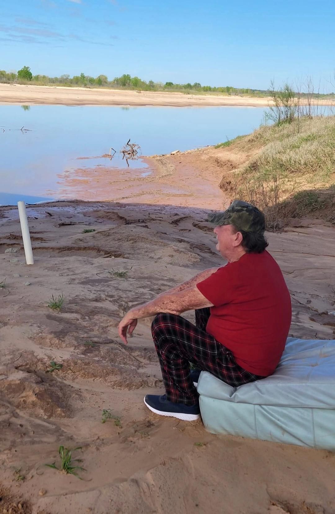Man in red shirt and plaid pants sits on mattress by a lake, looking at birds.