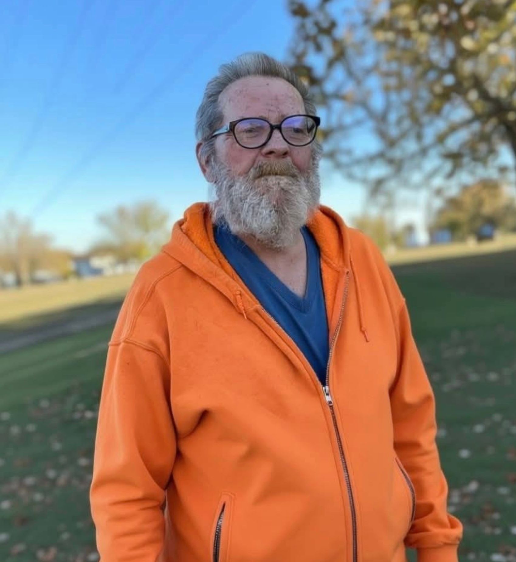 Man with gray beard wearing orange hoodie, standing outdoors under a blue sky.