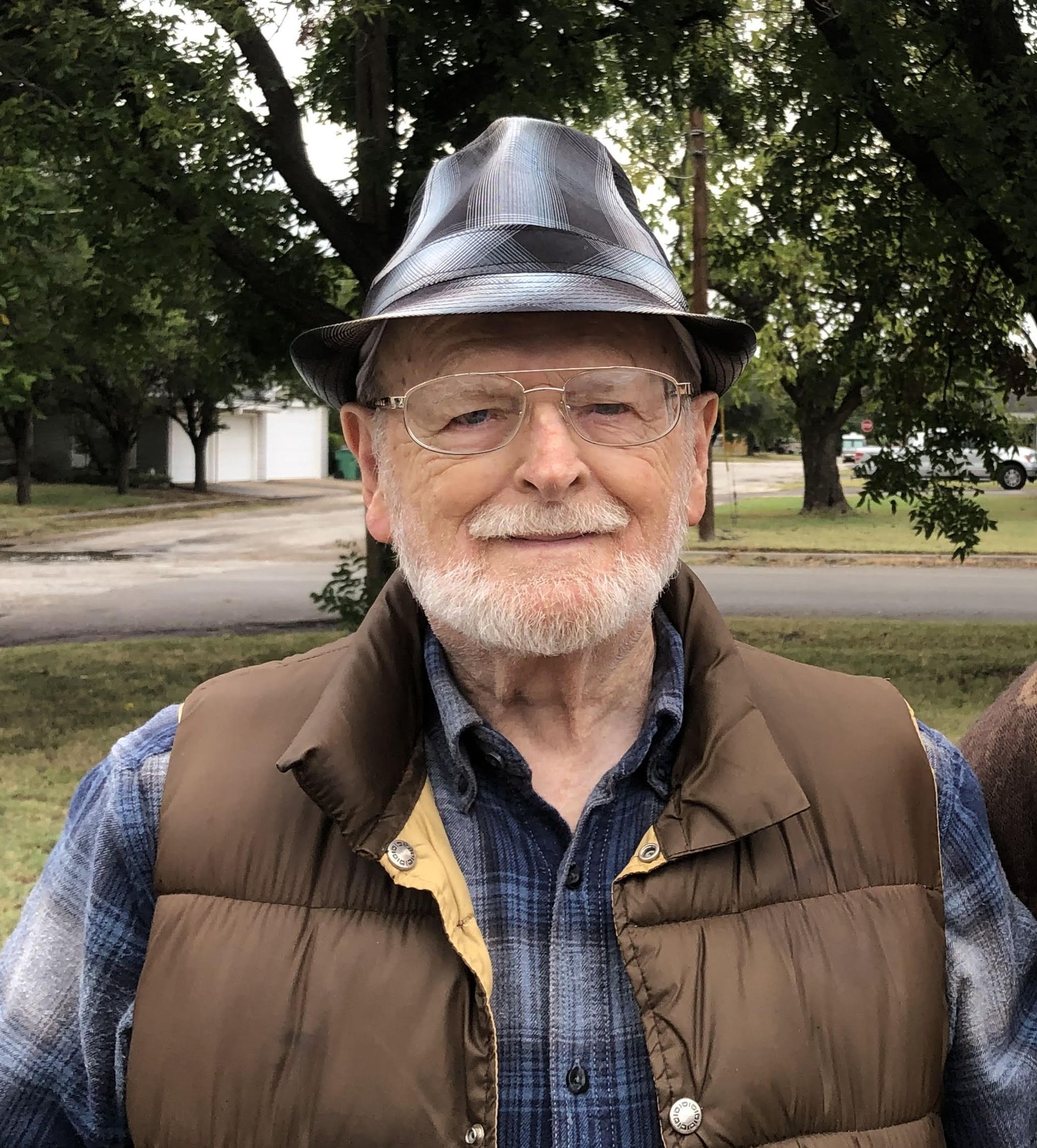 Older man wearing a hat, vest, and glasses, smiling in an outdoor setting.