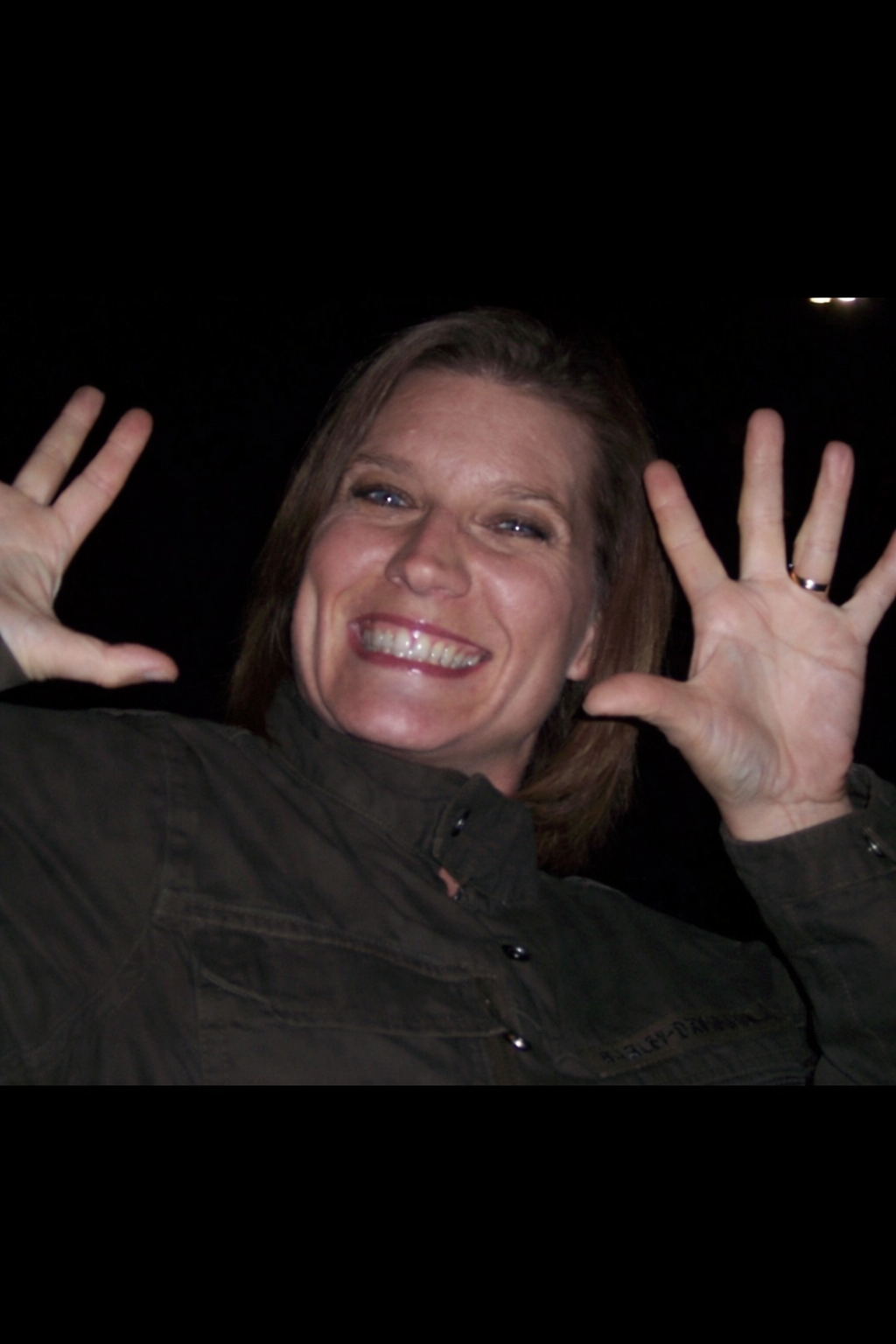 Woman with short brown hair smiles, holding up both hands, fingers spread wide. Dark background.