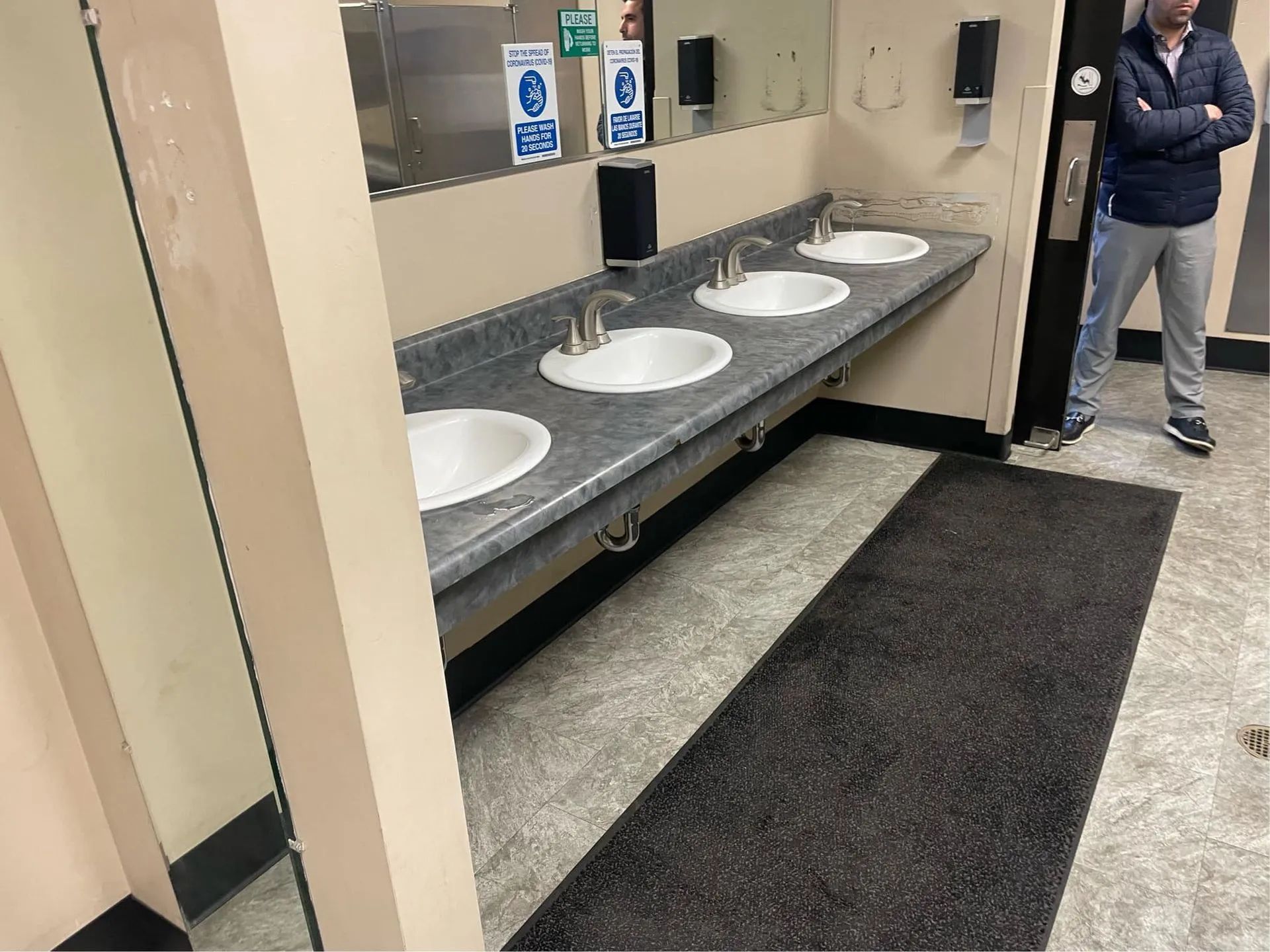 A man is standing in a bathroom with four sinks and a mirror.