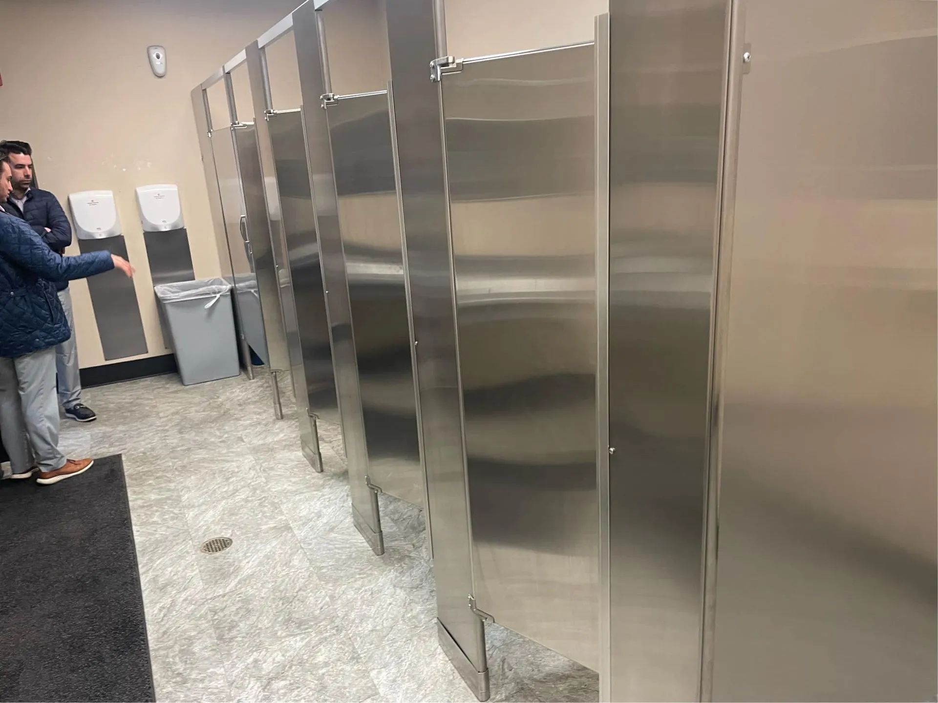 Two men are standing in a public restroom with stainless steel stalls.