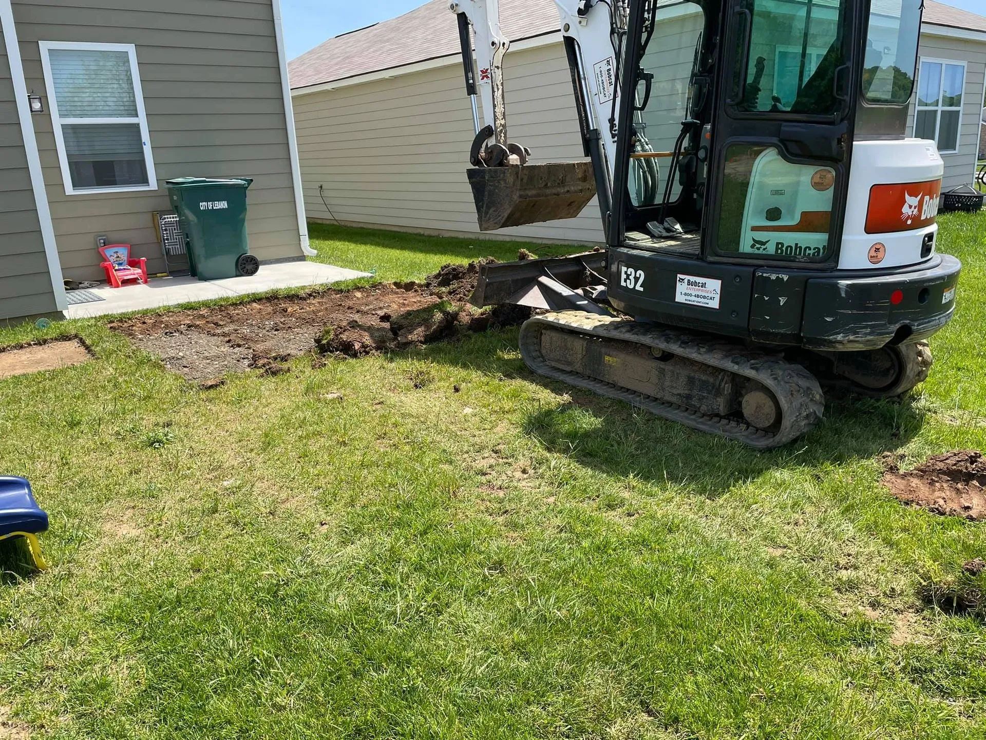 A small excavator is digging a hole in the grass in front of a house.