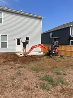 A man is standing in front of a house next to a small excavator.