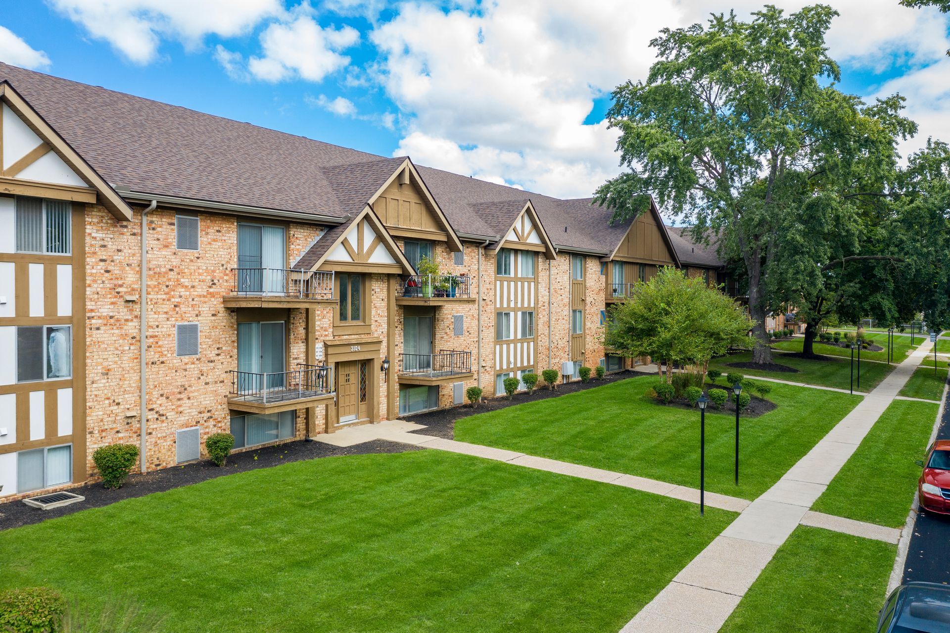 Apartment building with brick exterior, brown roof, and green lawn. Sidewalk and trees are present.