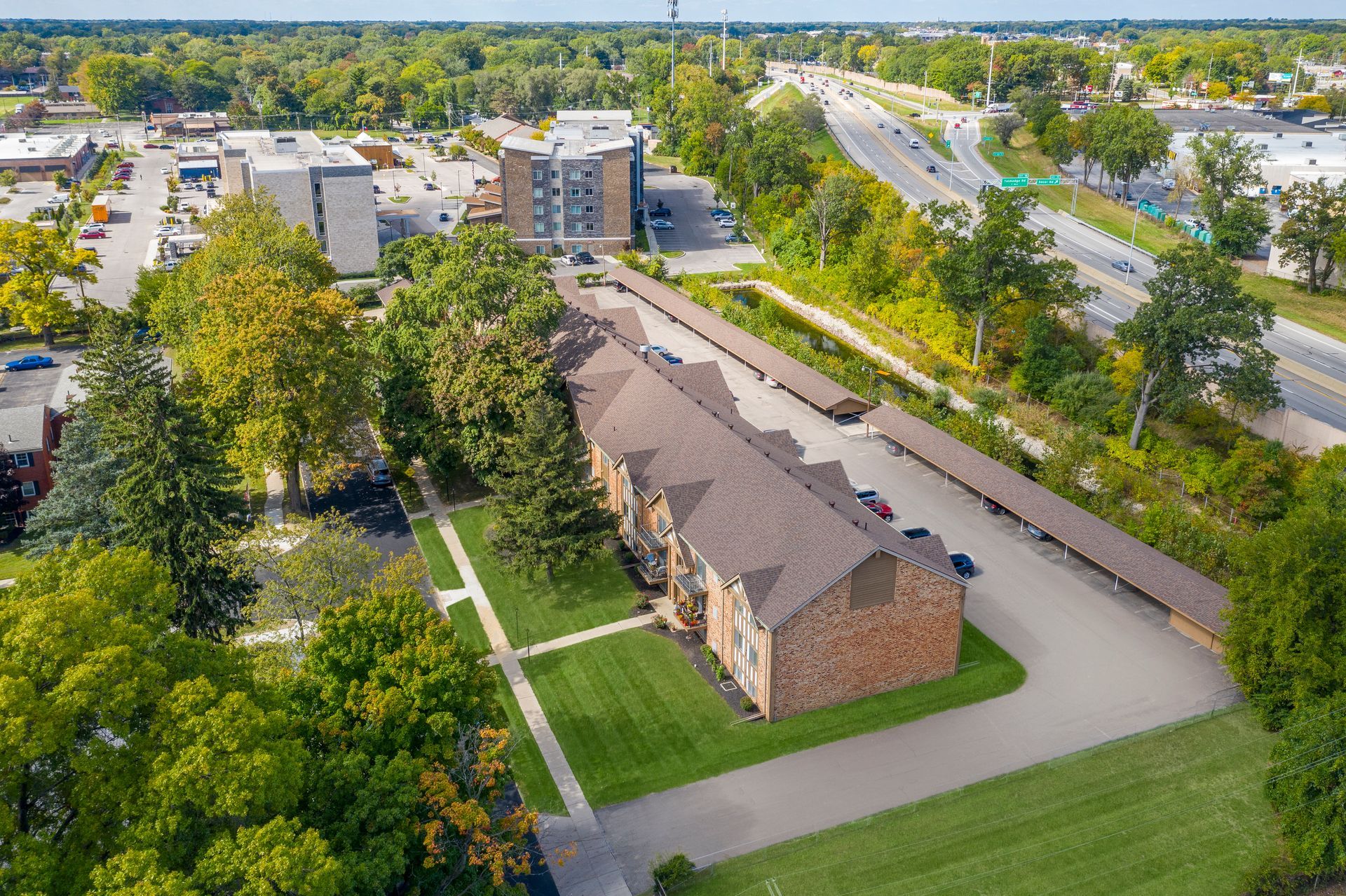 Aerial view of an apartment complex with a covered parking area, trees, and a road in the background.