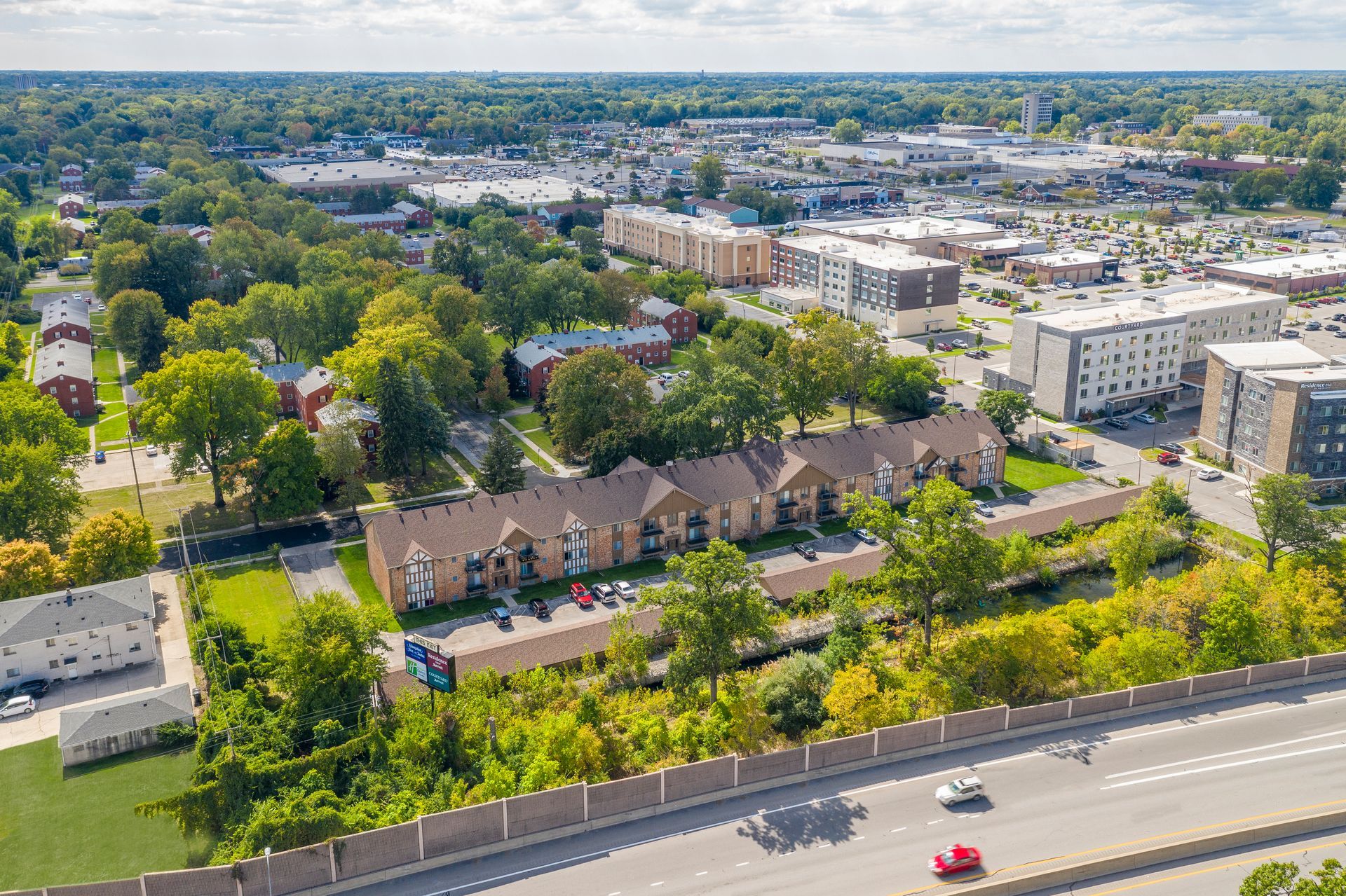 Aerial view of a suburban neighborhood with townhouses, trees, and commercial buildings. Cloudy sky.
