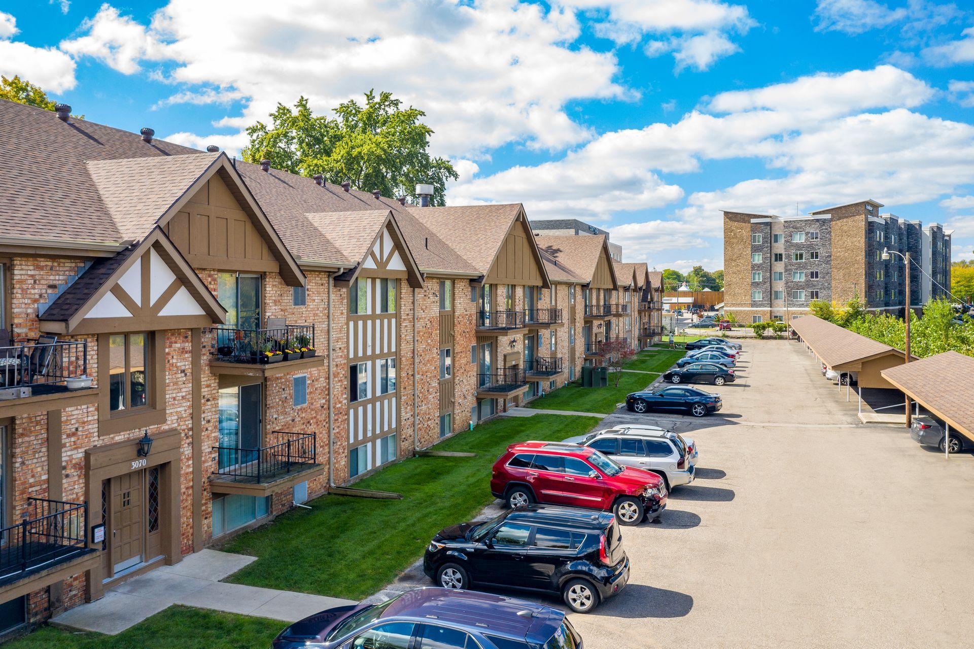 Apartment buildings with brown roofs and brick exteriors, cars parked in front, blue sky.