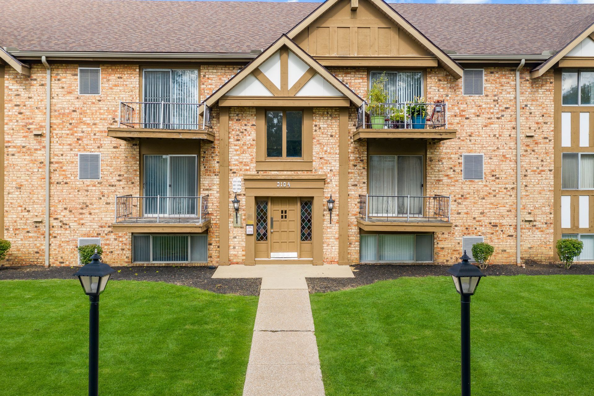 Apartment building exterior, brick and brown siding, with central entrance and balconies, green lawn, pathway.