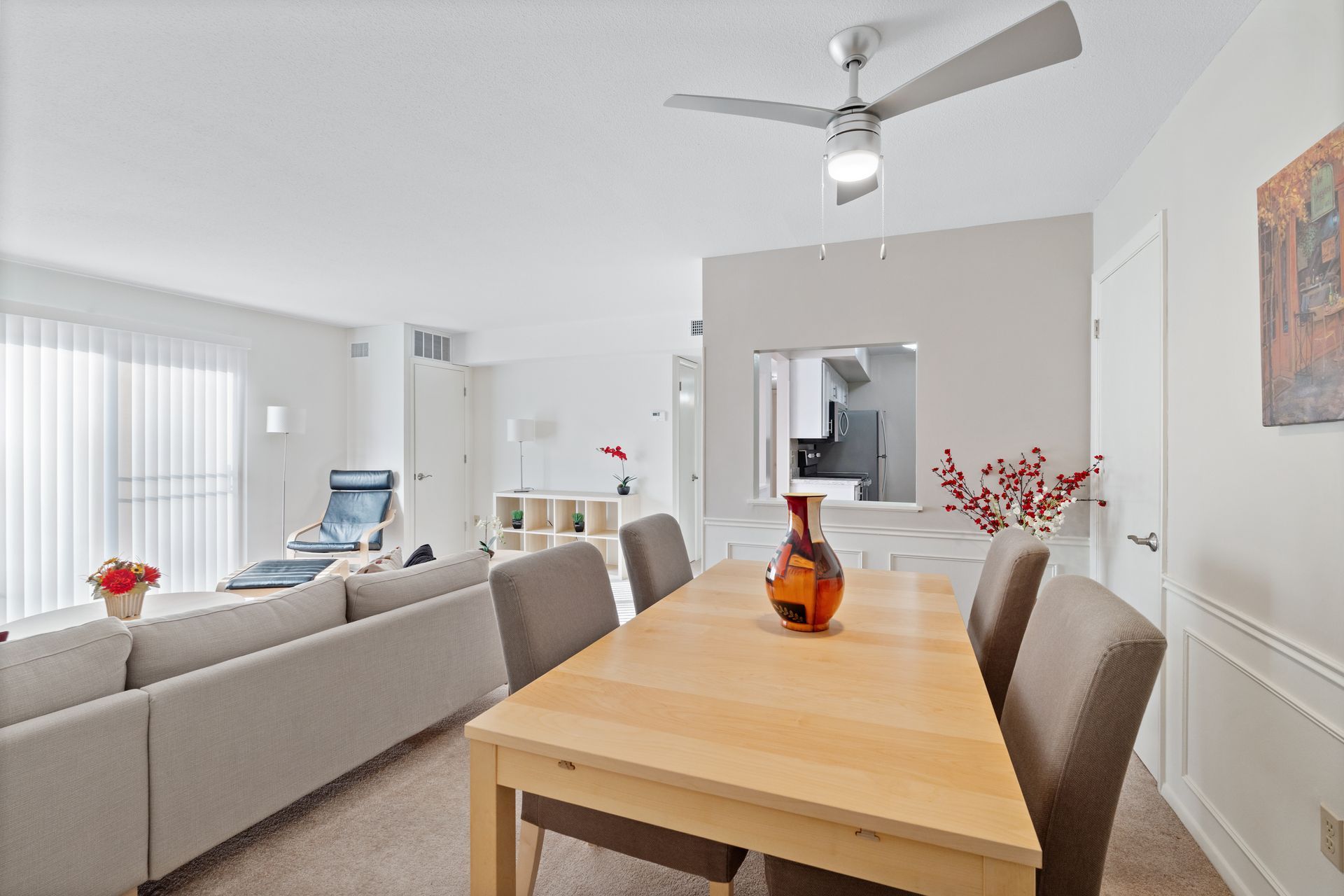 Dining area with table, chairs, vase, and view into kitchen, connecting to living room with couch and sliding door.