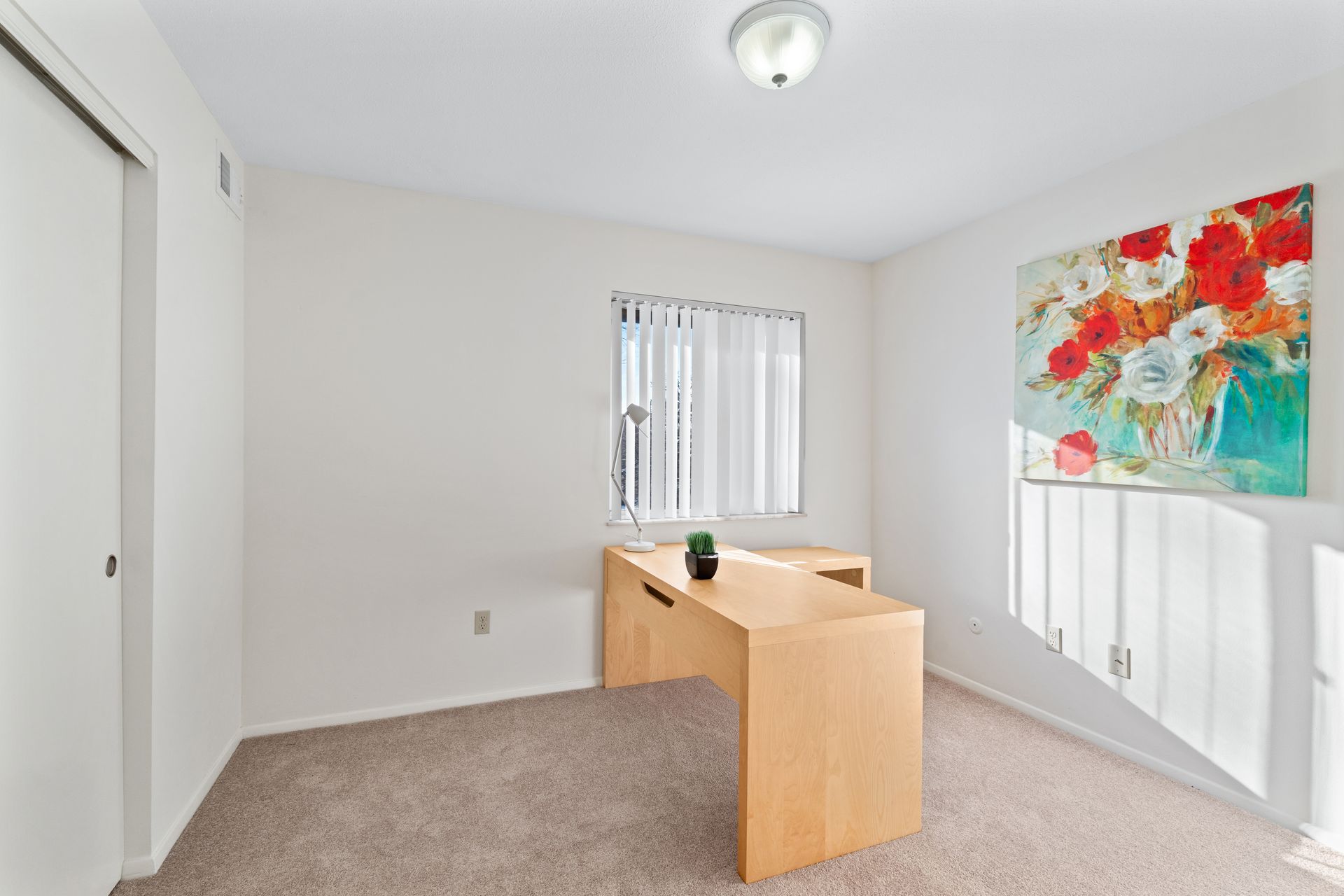 Light-filled home office with a modern wooden desk, a colorful painting, and a small window with blinds.