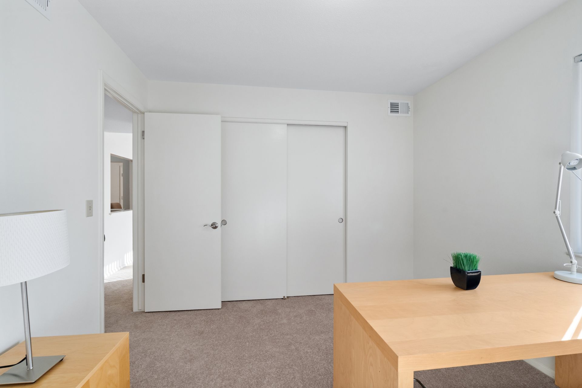 Empty office room with light wood desk, white walls, closet doors, and a small plant.