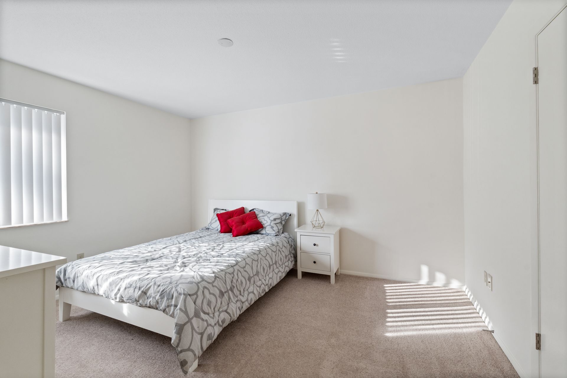 Bedroom with white walls, bed with patterned cover and red pillows, nightstand, and window with blinds.