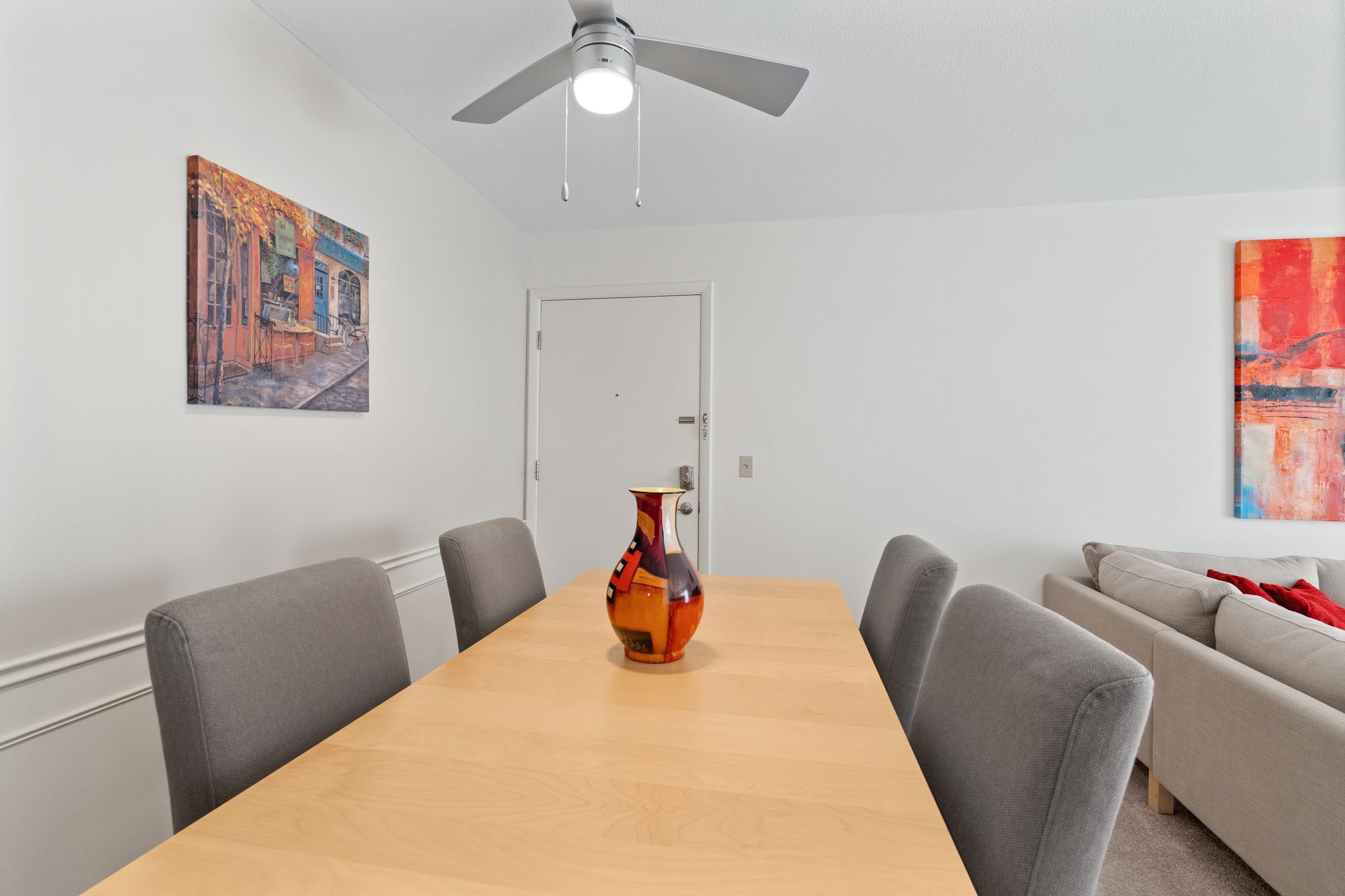 Dining room with wooden table, four gray chairs, vase, and wall art. White walls, ceiling fan, door.