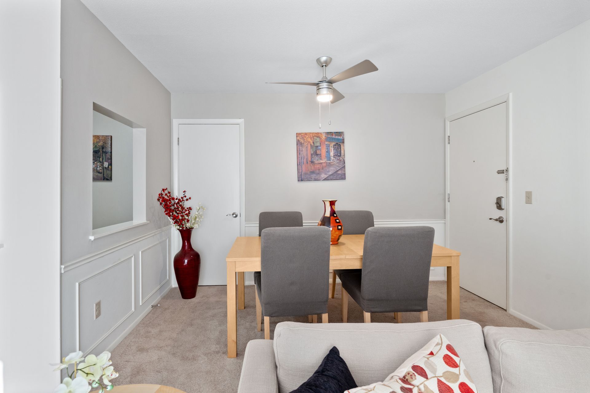 Dining area with wooden table, gray chairs, and red vase against a light gray wall.