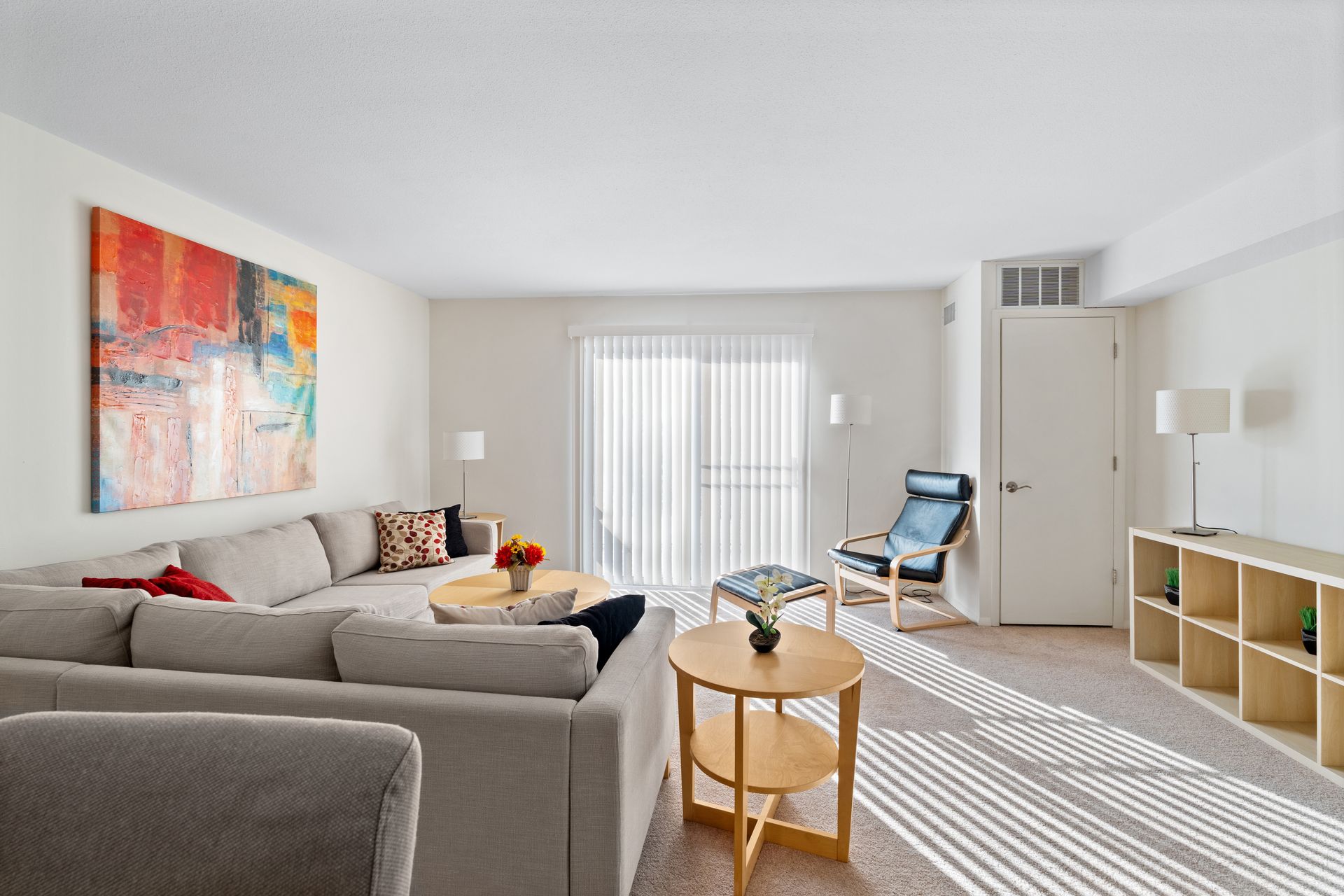 Bright living room with a large sectional sofa, colorful artwork, and a balcony door. Sunlight streams in.