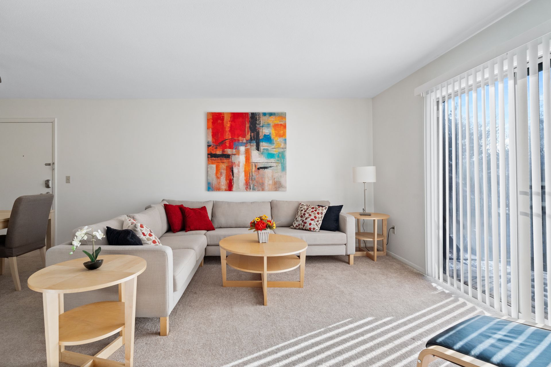 Bright living room with sectional sofa, coffee table, and colorful abstract artwork. Large window with blinds.