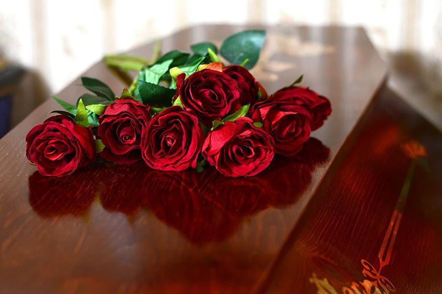 Red roses lie on a polished wooden casket, likely at a funeral.