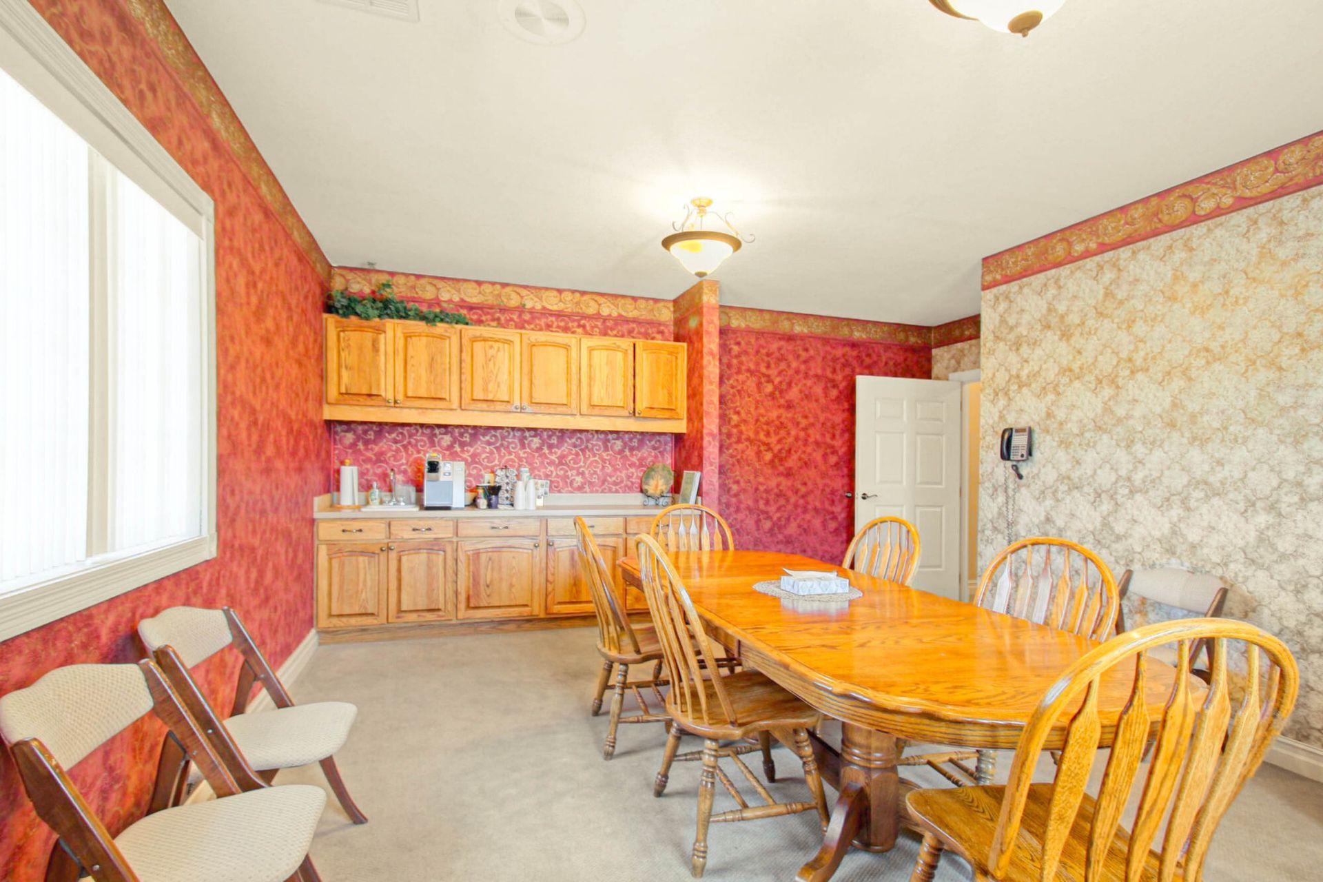 A dining room with wooden table, chairs, cabinets, and red and beige patterned wallpaper.