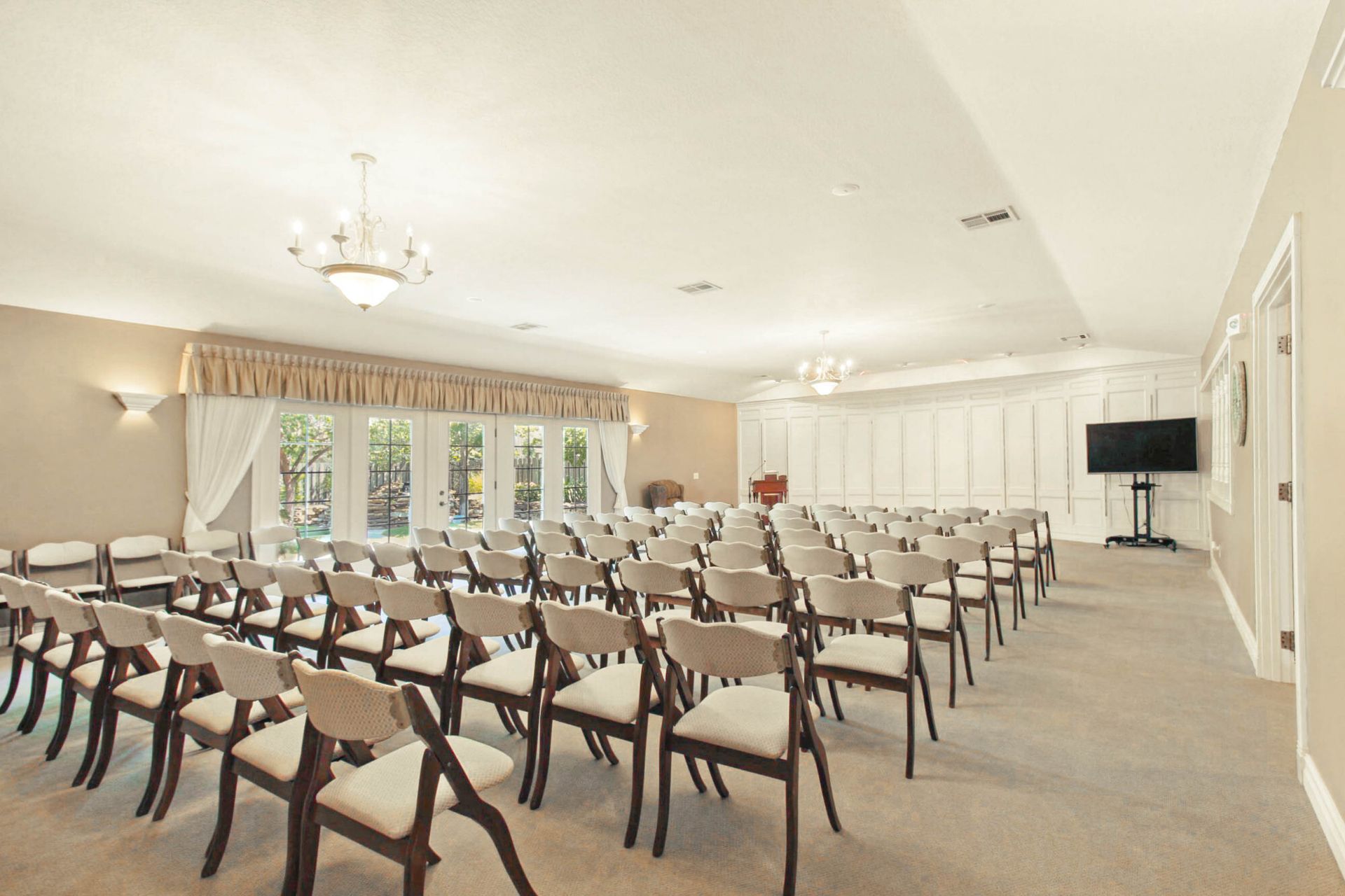 Rows of chairs face a stage in a light-filled room. A TV is on a stand; windows are behind a curtain.