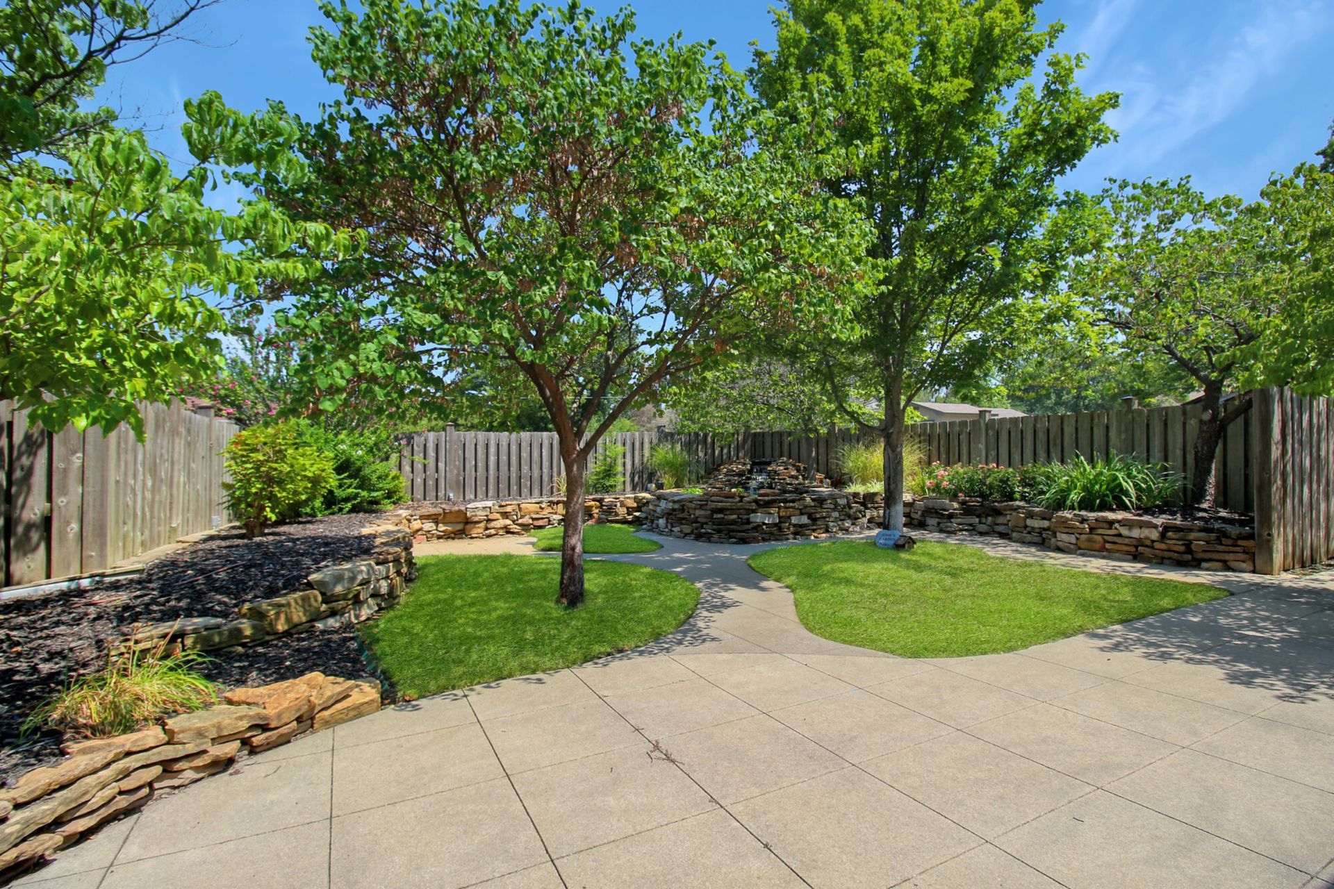 Patio in a backyard with trees, grass, and a wooden fence.