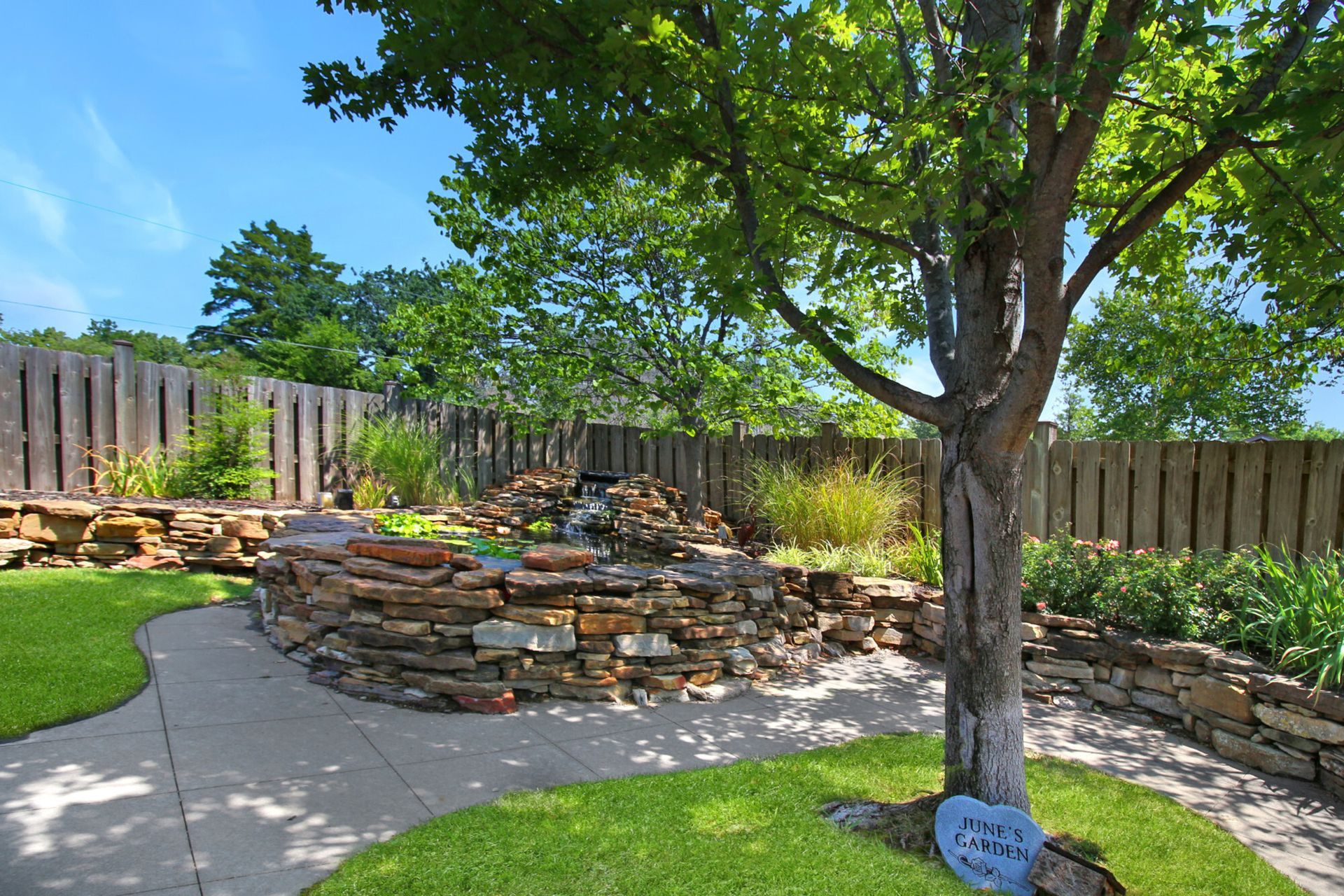 Stone-walled pond with small waterfall, path, tree, and fence on a sunny day.