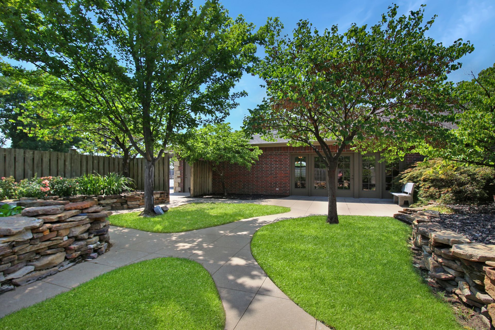 Green lawn with curved walkway leading to a brick building, bordered by trees and stone walls.
