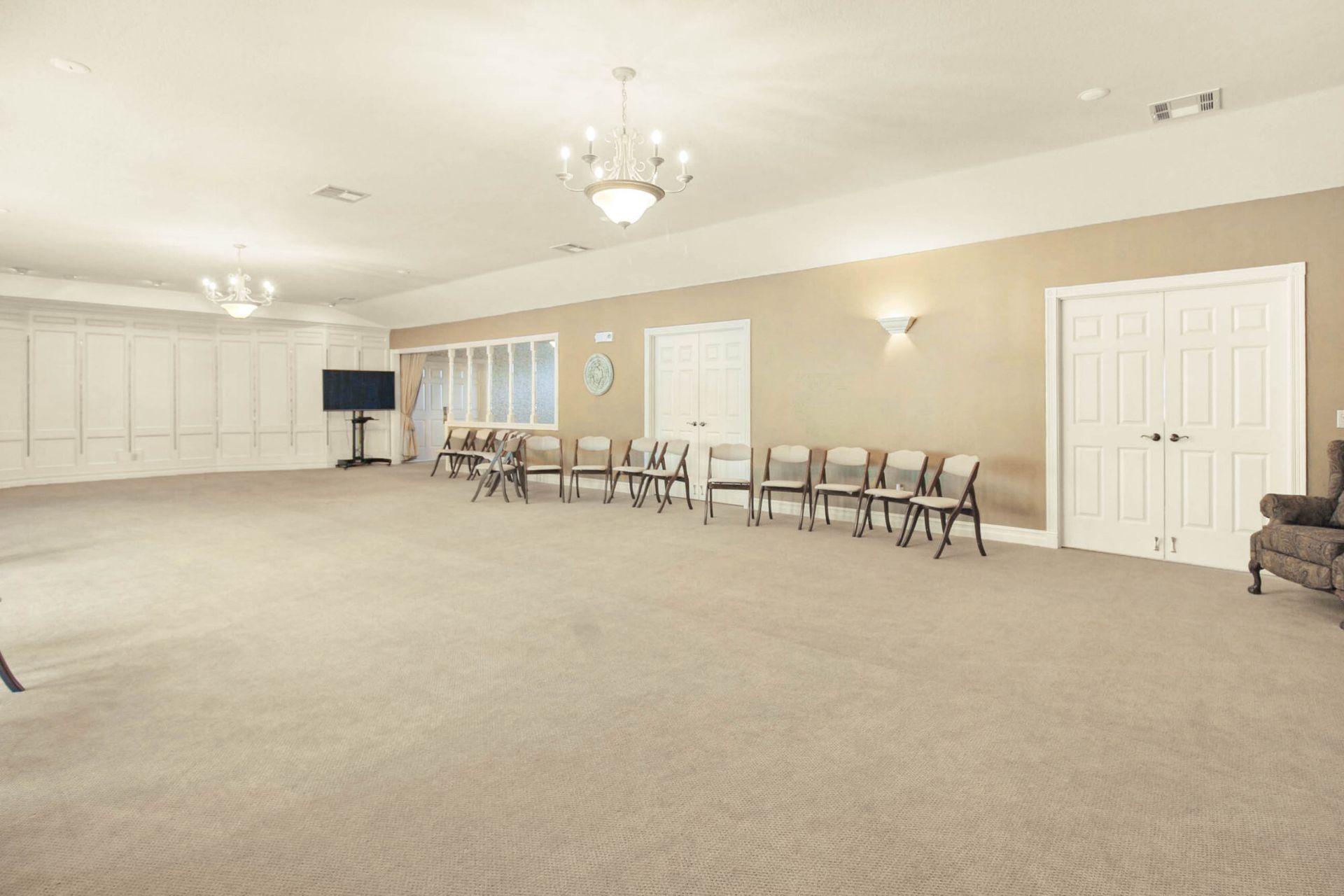 Empty event space with beige walls, carpet, chairs, and two chandeliers.