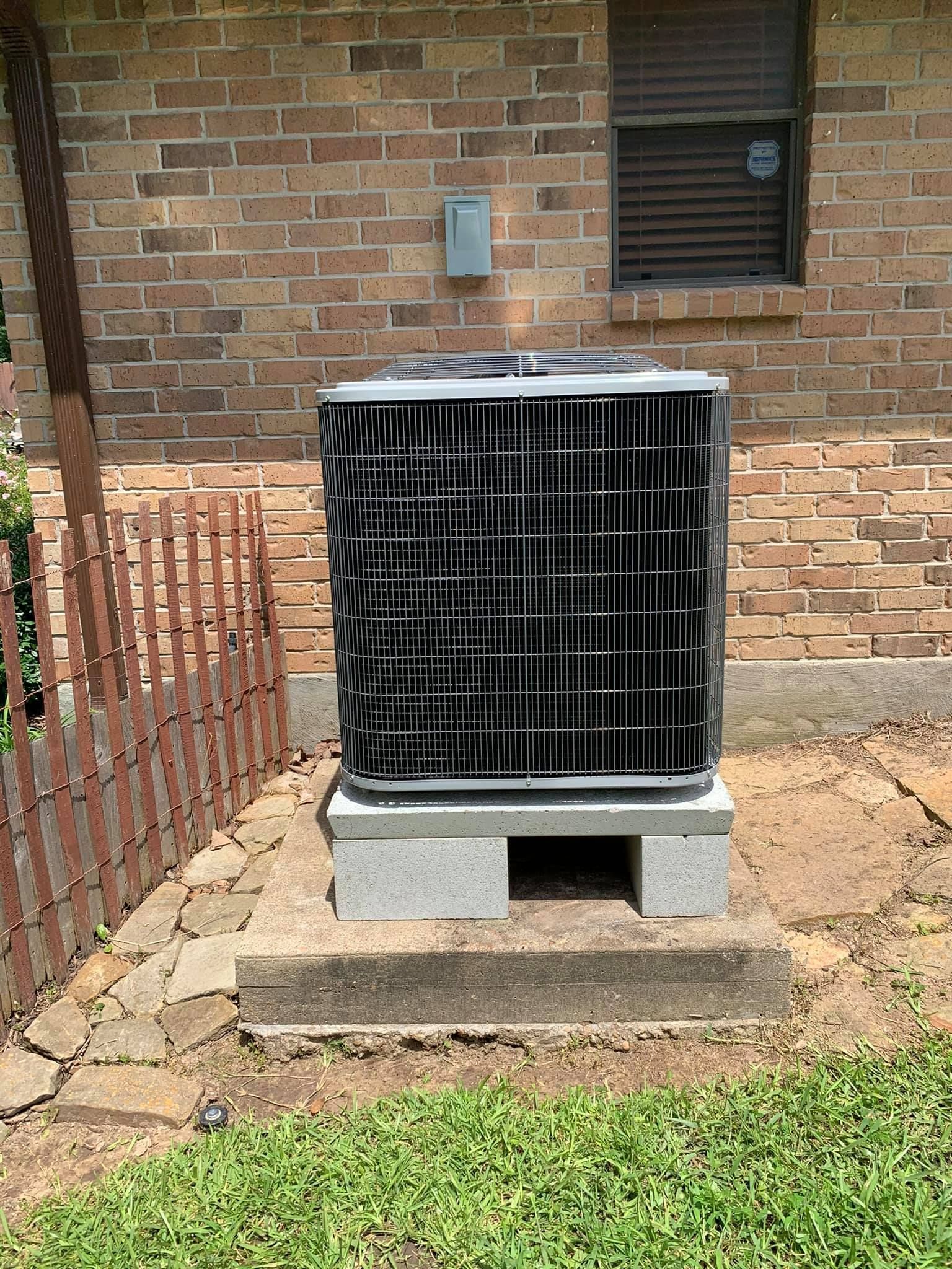 An air conditioner is sitting on top of a concrete block in front of a brick building.