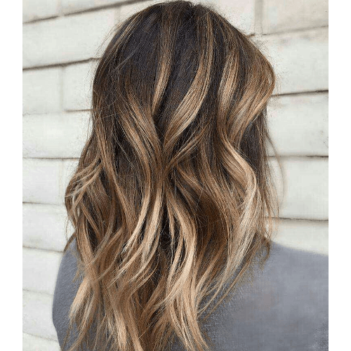 A woman with long wavy brown hair is standing in front of a white wall.