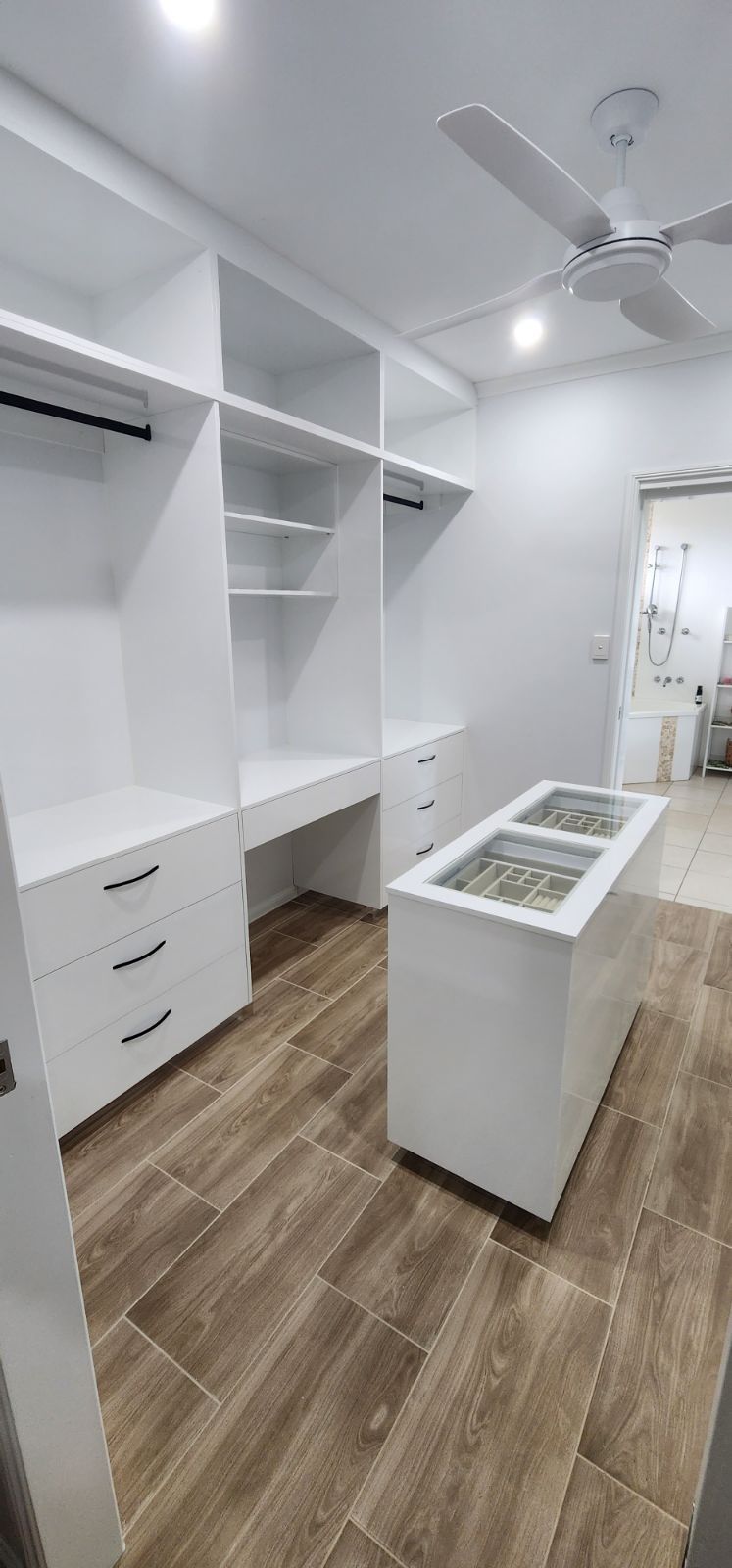 A white walk in wardrobe with a bathroom in the background and a wooden floor— Cairns Custom Wardrobes in Manunda, QLD