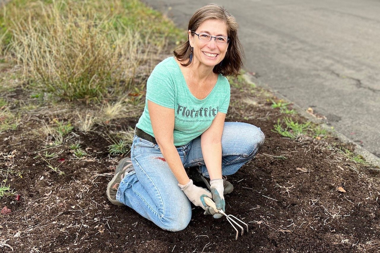 A woman is kneeling down in the dirt holding a rake.