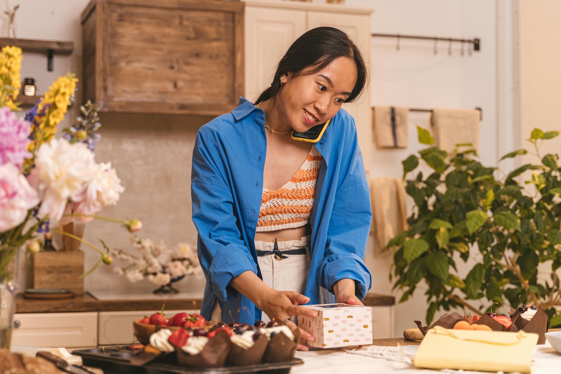 A woman is cutting a cake in a kitchen while talking on a cell phone.