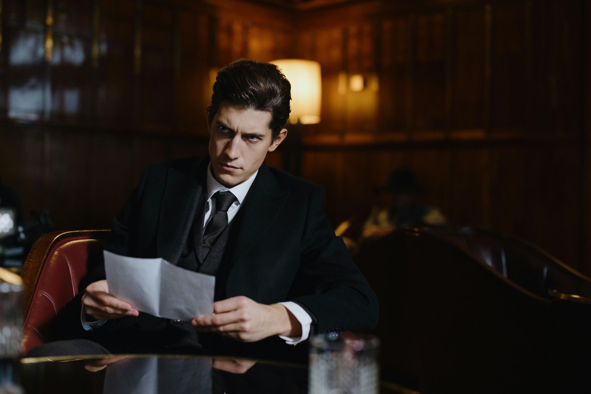 A man in a suit and tie is sitting at a table reading a piece of paper.