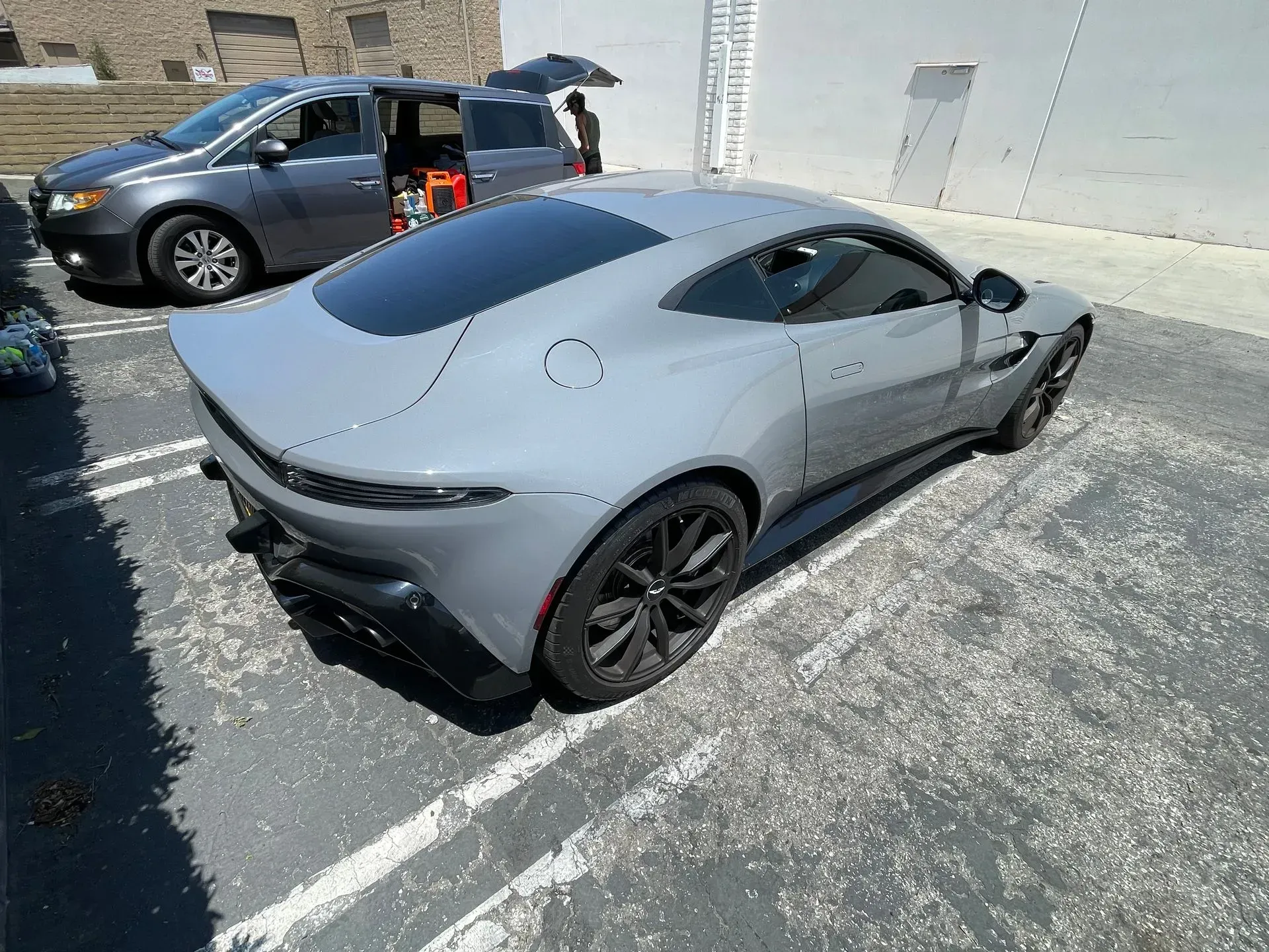 A high-angle view of a grey Aston Martin sports car parked in an outdoor lot next to a grey minivan.