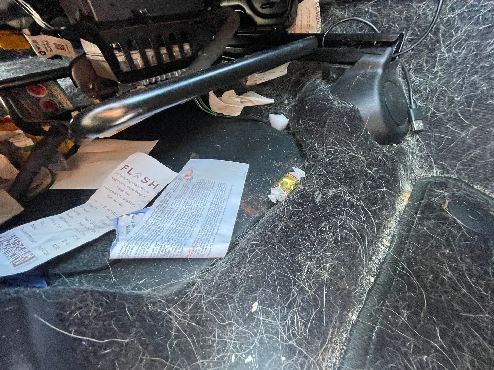 A vehicle floor covered in pet hair, with two pieces of crumpled paper and a small plastic tube near the seat rails.