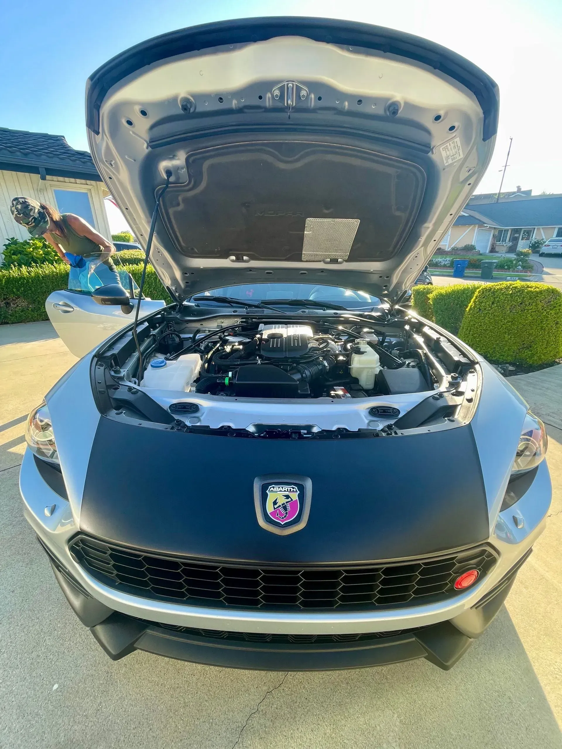 A silver car with its hood open, parked on a driveway with a person visible inside the open driver-side door.