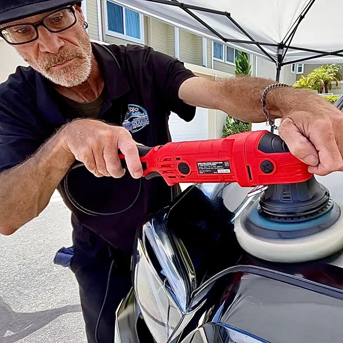 A person in a black shirt uses a red power buffer to polish the glossy black hood of a car.