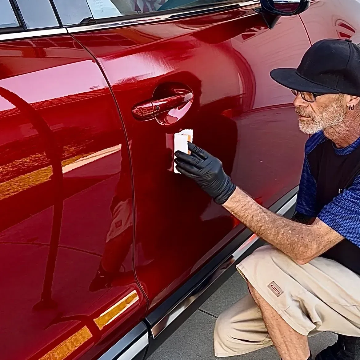 A person in gloves and a baseball cap applies a white touch-up tool to a scratch on a shiny red car door.