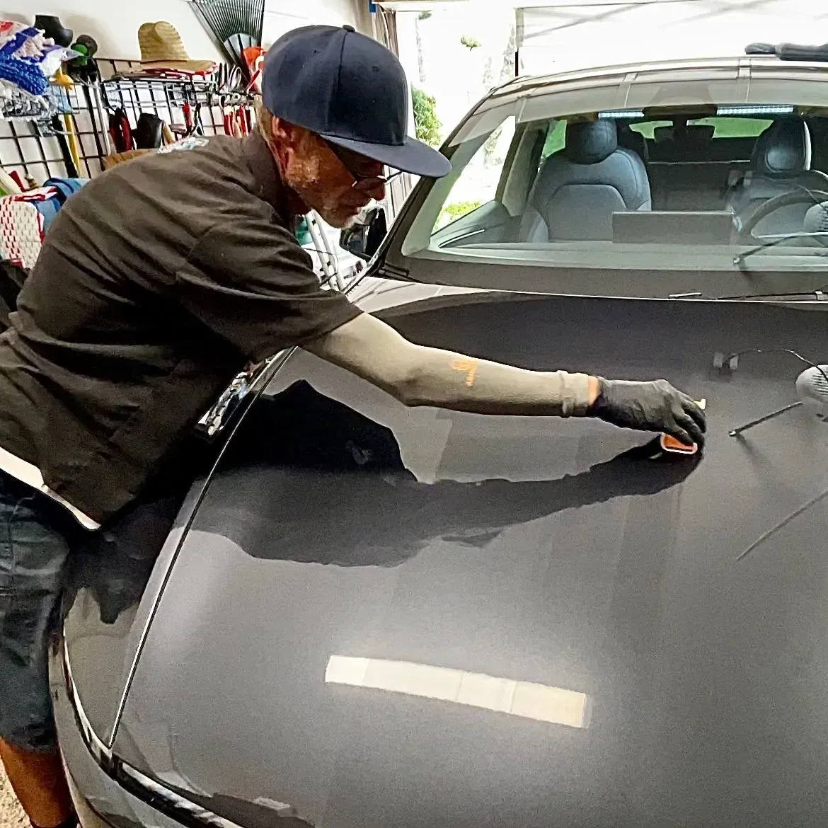 A person wearing a dark shirt and hat applies a product to a car hood with a microfiber cloth in a garage.