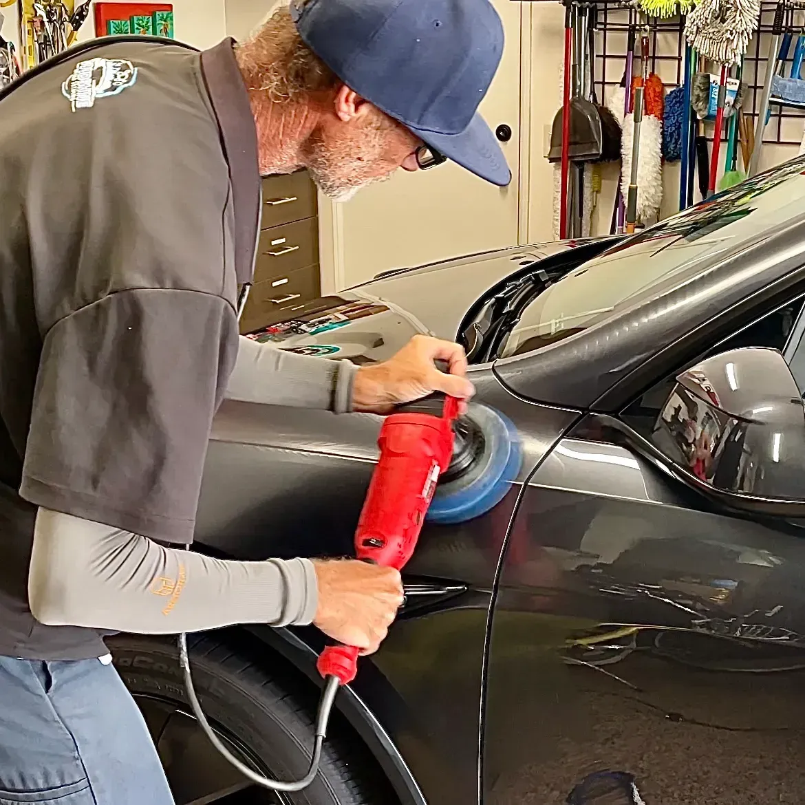 A person wearing a blue cap polishes the hood of a dark car with a red handheld electric buffer in a garage.